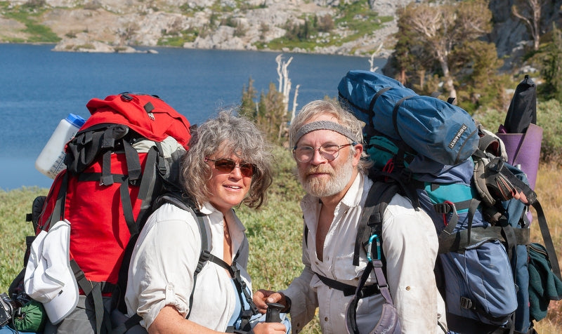 A woman and man with heavy back packs  and grey hair hiking by a lake.